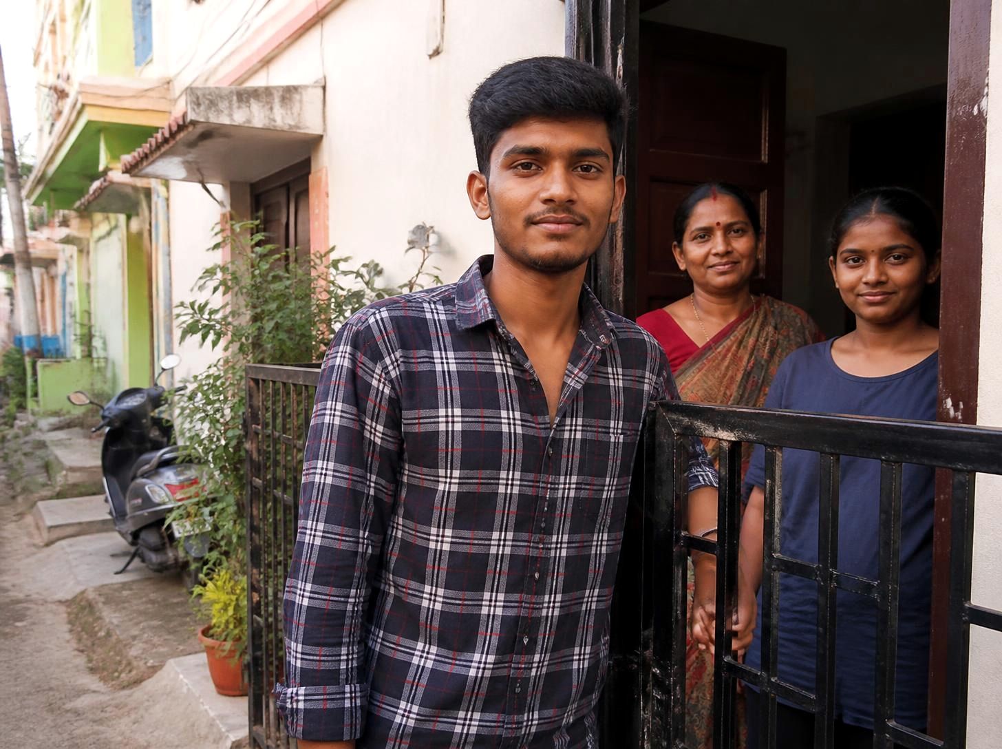 Young man at the gate of his family home in a small-town lane, his mother and sister in the doorway